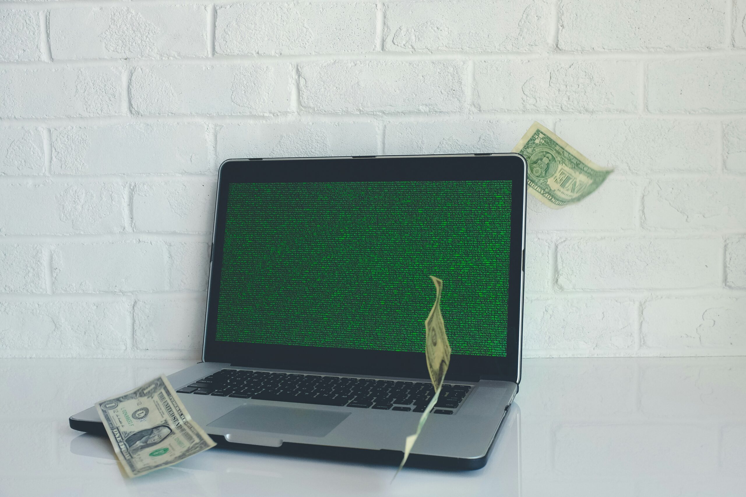 A laptop on a desk, with dollar notes dropping beside it